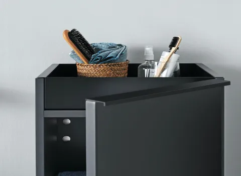 Ein geöffneter, schwarzer burgbad Fiumo Schrank zeigt Pflegeprodukte wie Zahnbürsten, eine Haarbürste und Handtücher, die ordentlich in einem kleinen Korb verstaut sind.