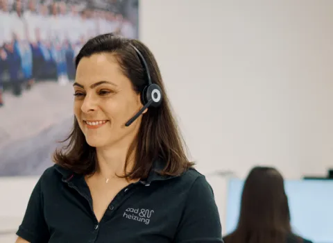 Eine Frau mit einem Headset sitzt lächelnd an einem Schreibtisch in einem modernen Büro.