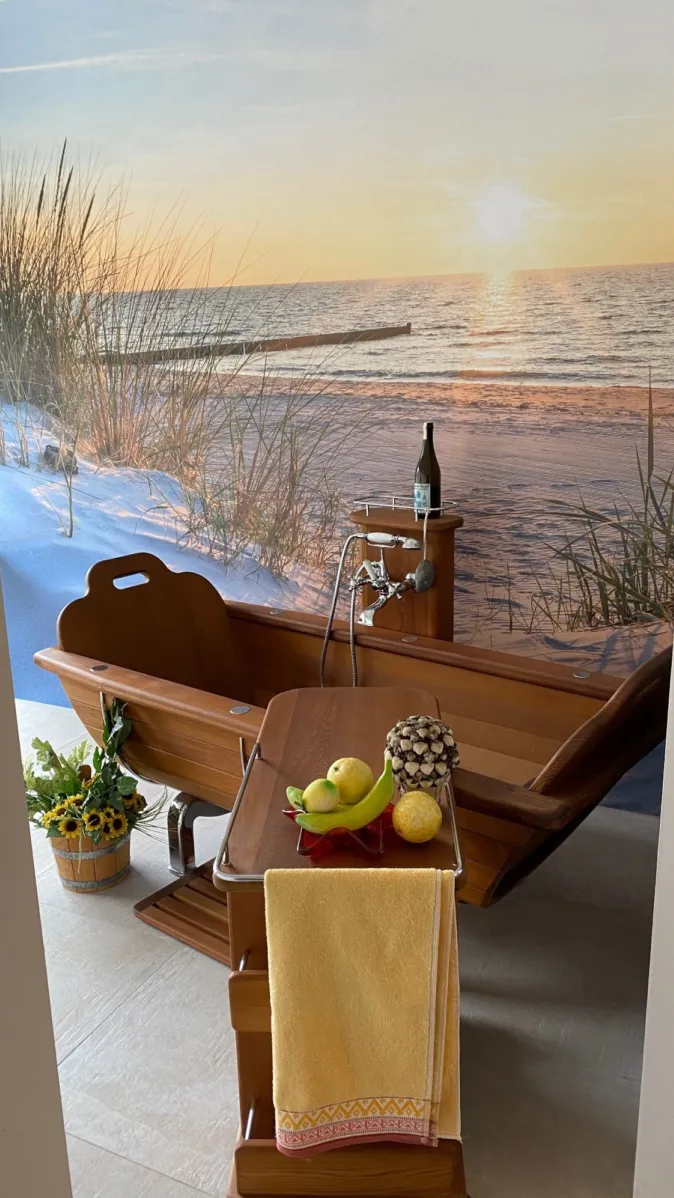 Eine nostalgisch anmutende Holzbadewanne steht in einem Raum mit Strandtapete und Meerblick im Sonnenuntergang.