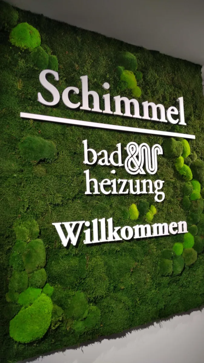 Eine dekorative Mooswand mit kräftig grüner Struktur trägt den weißen Schriftzug „Schimmel – bad & heizung – Willkommen“.