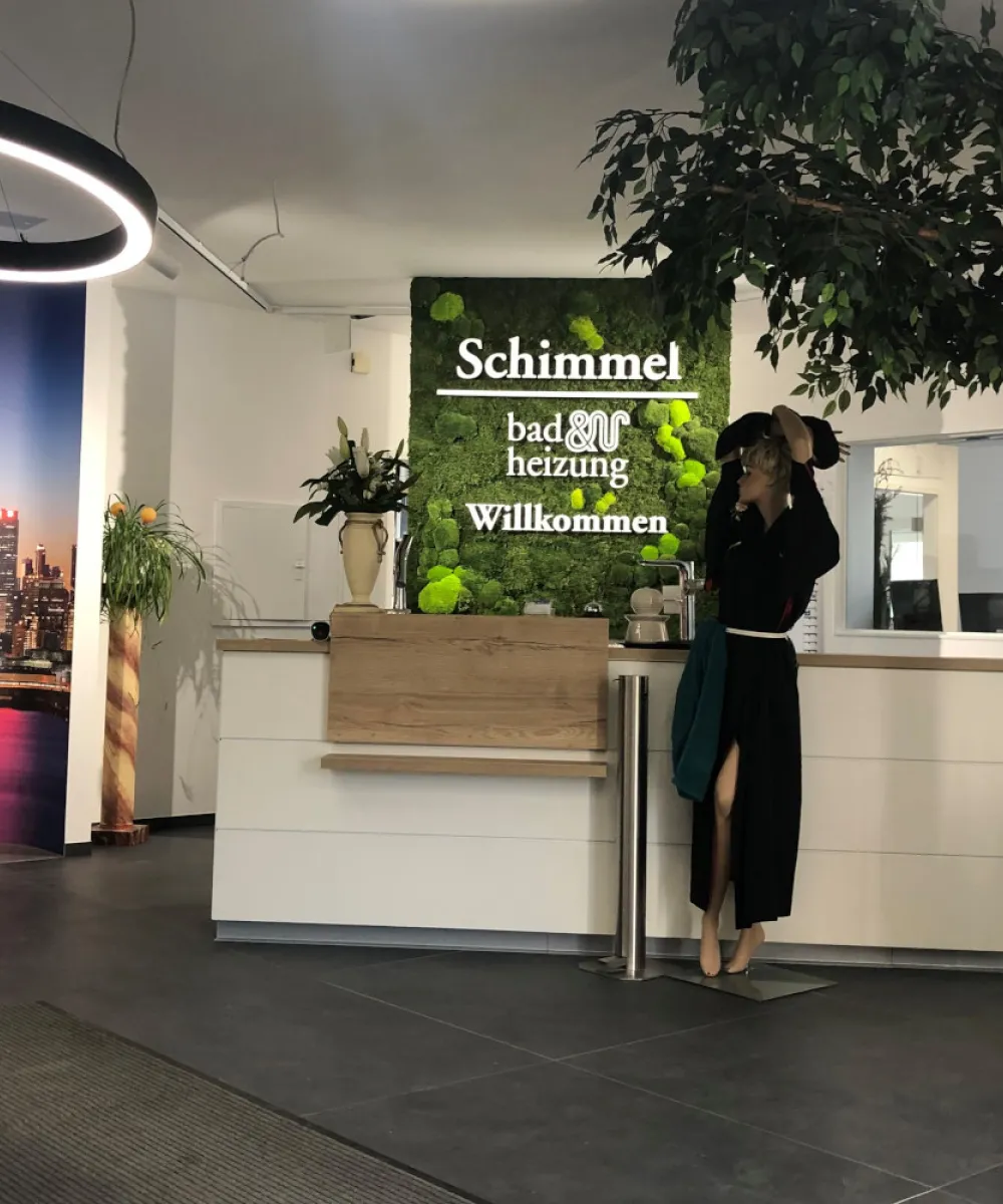 Ein freundlicher Empfangsbereich mit Mooswand-Schriftzug und heller Theke wird durch große Pflanzen und ein dekoratives Stadtbild aufgelockert.