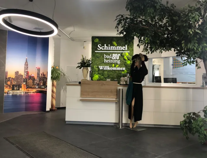 Ein freundlicher Empfangsbereich mit Mooswand-Schriftzug und heller Theke wird durch große Pflanzen und ein dekoratives Stadtbild aufgelockert.