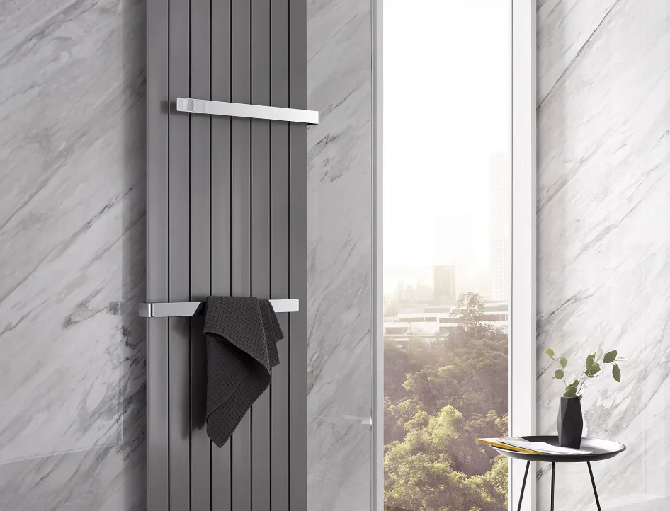 Ein vertikaler HSK Designheizkörper in Grau, an einer eleganten Marmowand montiert, mit Handtuch und Ausblick durch ein großes Fenster.
